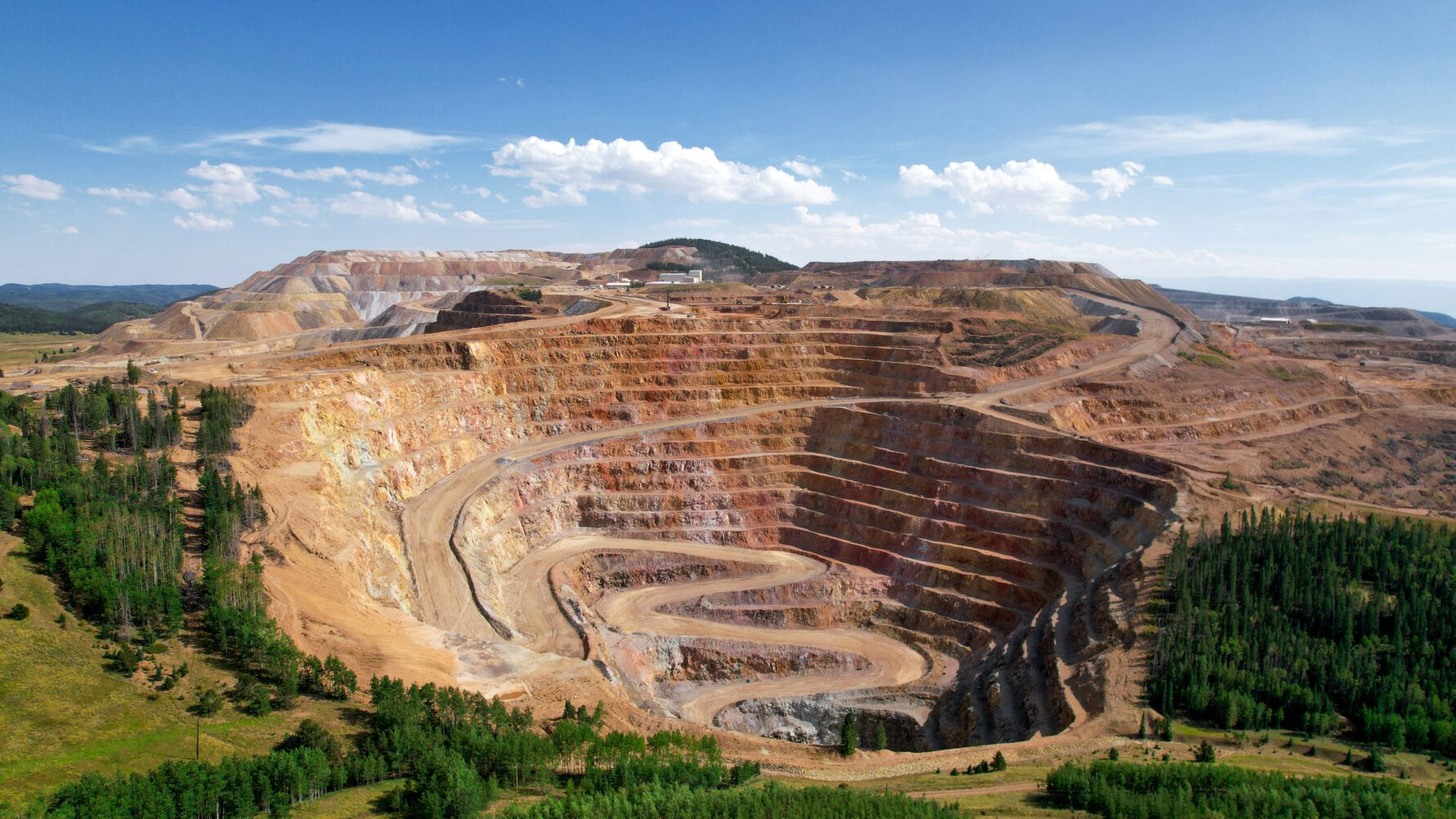 A wide aerial shot of a massive open-pit terrace mine with spiraling dirt roads leading to the center, surrounded by green forests under a clear blue sky.