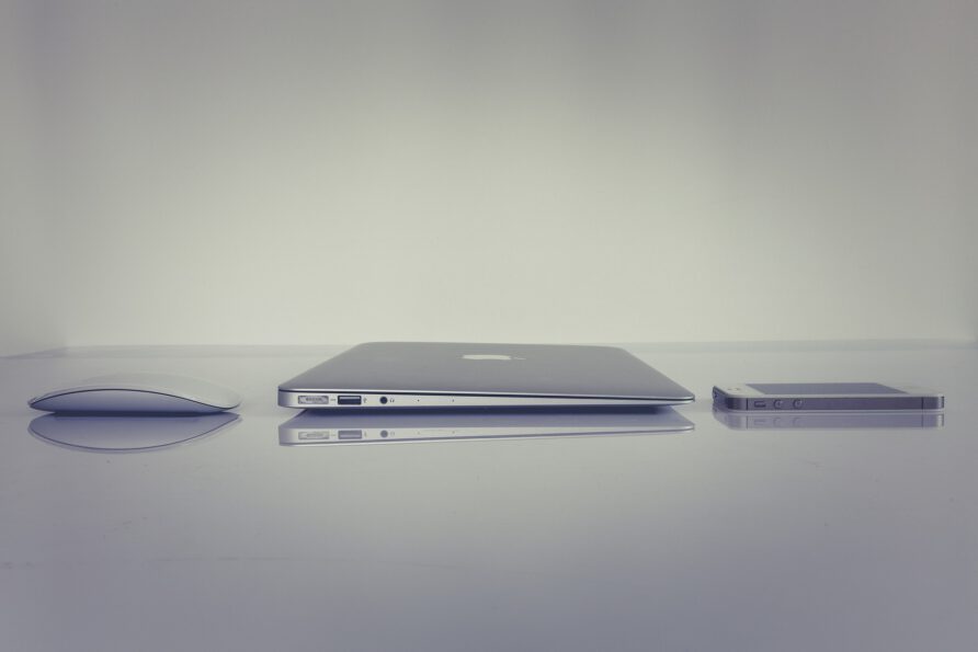 A side profile view of a silver Apple laptop, an iPhone, and a Magic Mouse arranged in a row on a reflective white surface against a plain white background.