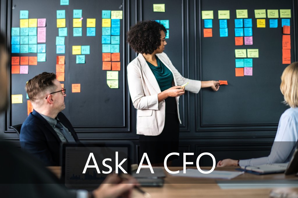 A professional woman in a white blazer stands at a black wall covered in colorful sticky notes, pointing to a specific note while leading a meeting with two colleagues seated at a conference table. Overlay text at the bottom reads Ask A CFO.