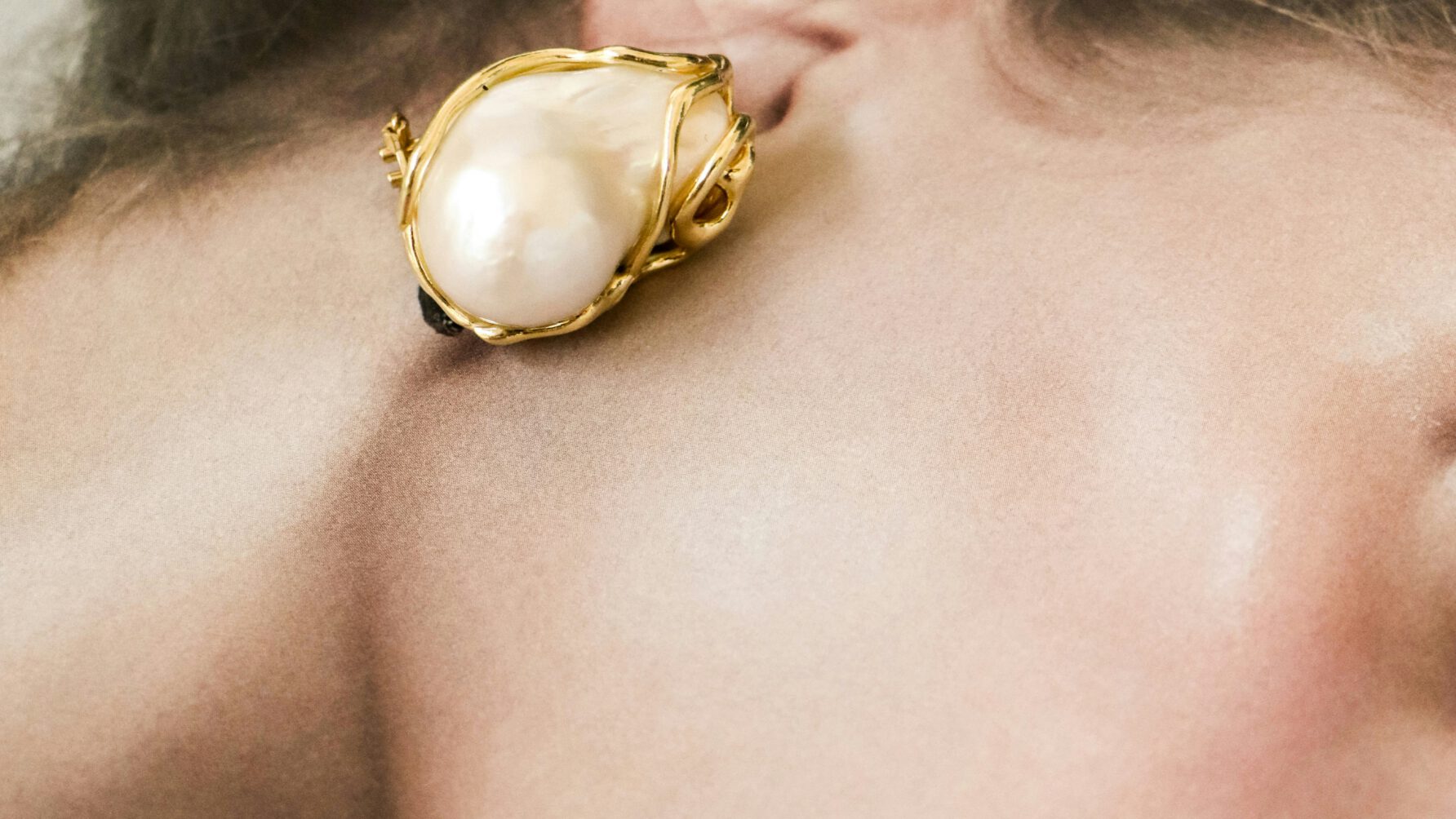 A close-up shot of a woman with her hair in a bun wearing a large, organic-shaped baroque pearl earring encased in a textured gold setting.