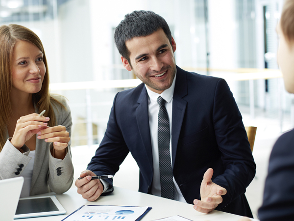 A diverse group of business professionals in formal attire engaging in a positive and productive discussion during an office meeting, with data charts and a tablet visible on the table.