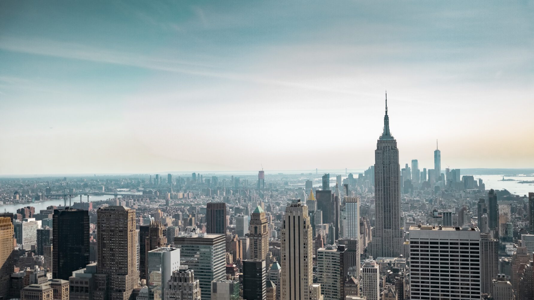 A high-angle, wide-lens view of the New York City skyline featuring the Empire State Building prominently in the foreground under a hazy blue sky.