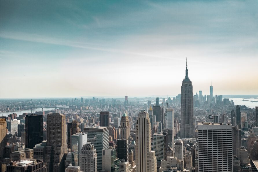 A high-angle, wide-lens view of the New York City skyline featuring the Empire State Building prominently in the foreground under a hazy blue sky.