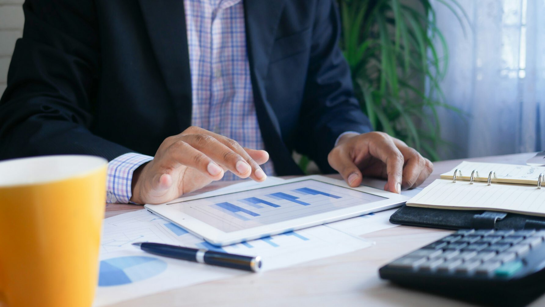 A person in a dark blazer and checkered shirt reviews blue bar charts on a white tablet at a desk equipped with a calculator, notebook, and a yellow coffee mug.