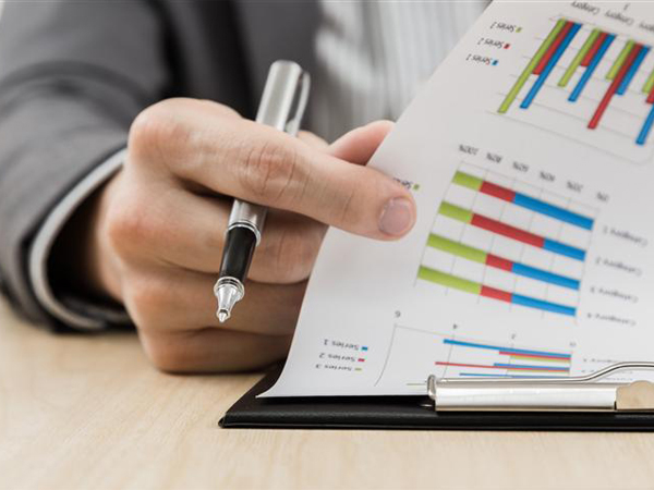 A close-up view of a professional's hand holding a silver pen while reviewing printed financial reports featuring colorful bar graphs and data analytics on a clipboard.
