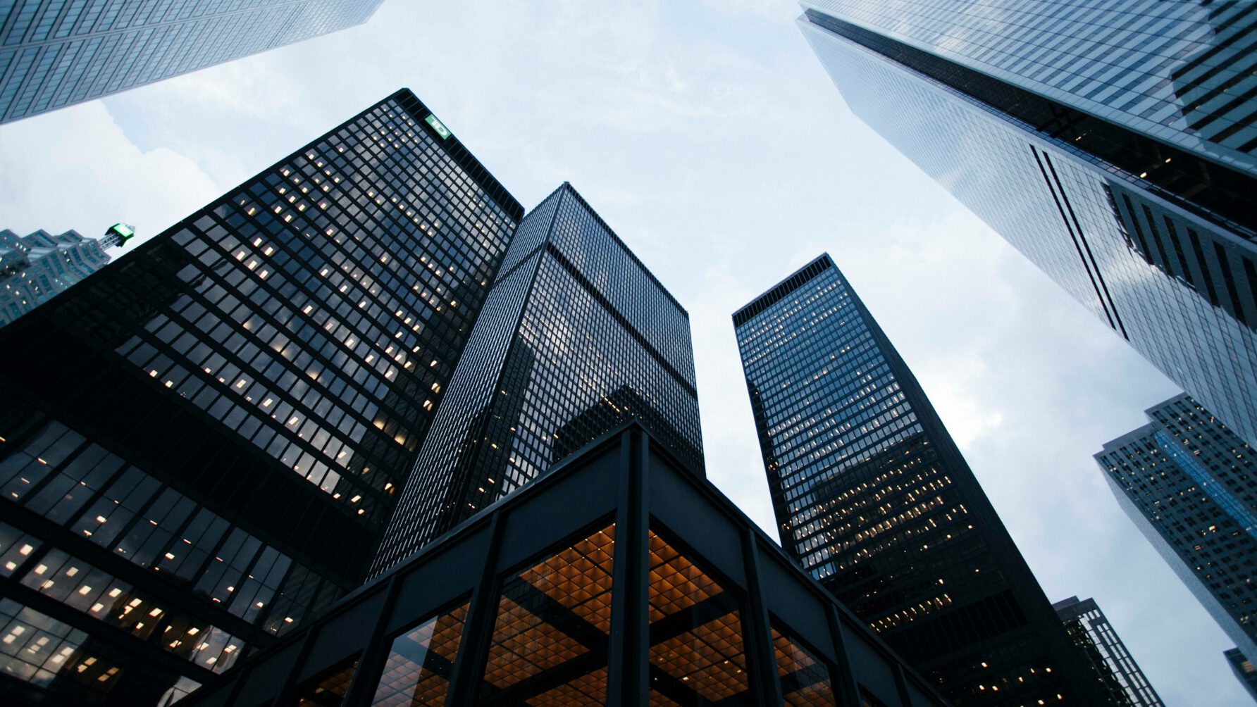 A dramatic low-angle shot looking up at several dark glass and steel corporate skyscrapers reaching toward a cloudy sky, with glowing office lights visible through the windows.