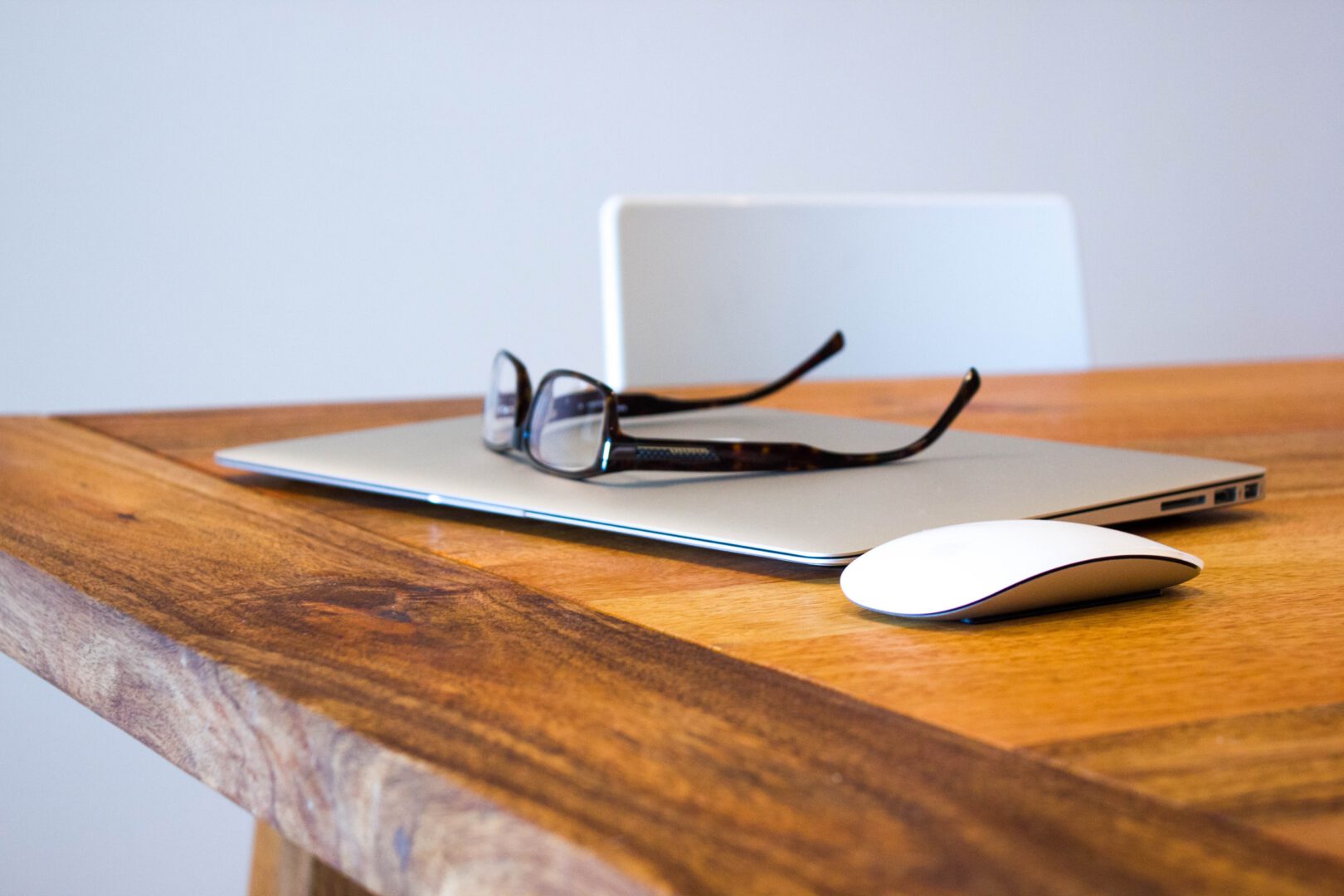 A close-up, side-profile view of a minimalist workspace on a natural wooden table. A pair of tortoiseshell glasses rests on top of a closed silver laptop next to a white wireless mouse.