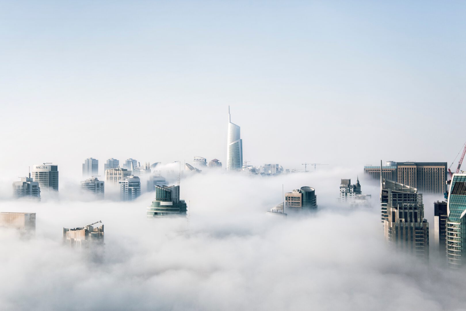 A breathtaking high-angle shot of a metropolitan skyline with modern skyscrapers piercing through a dense, white layer of morning fog and low-lying clouds under a clear, bright sky.