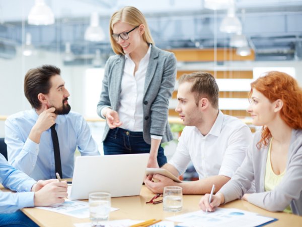 A team of four professionals collaborating around a light wood desk in a bright, modern office; one woman stands while leading the discussion as her colleagues look on, using laptops and tablets.
