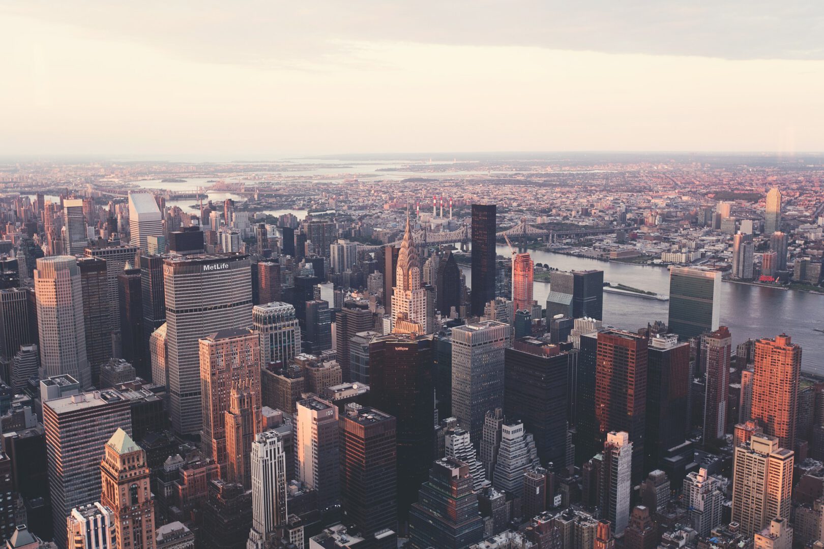 An expansive aerial view of the New York City skyline during sunset, showing the Chrysler Building and other Manhattan skyscrapers bathed in warm light, overlooking the East River and distant bridges.