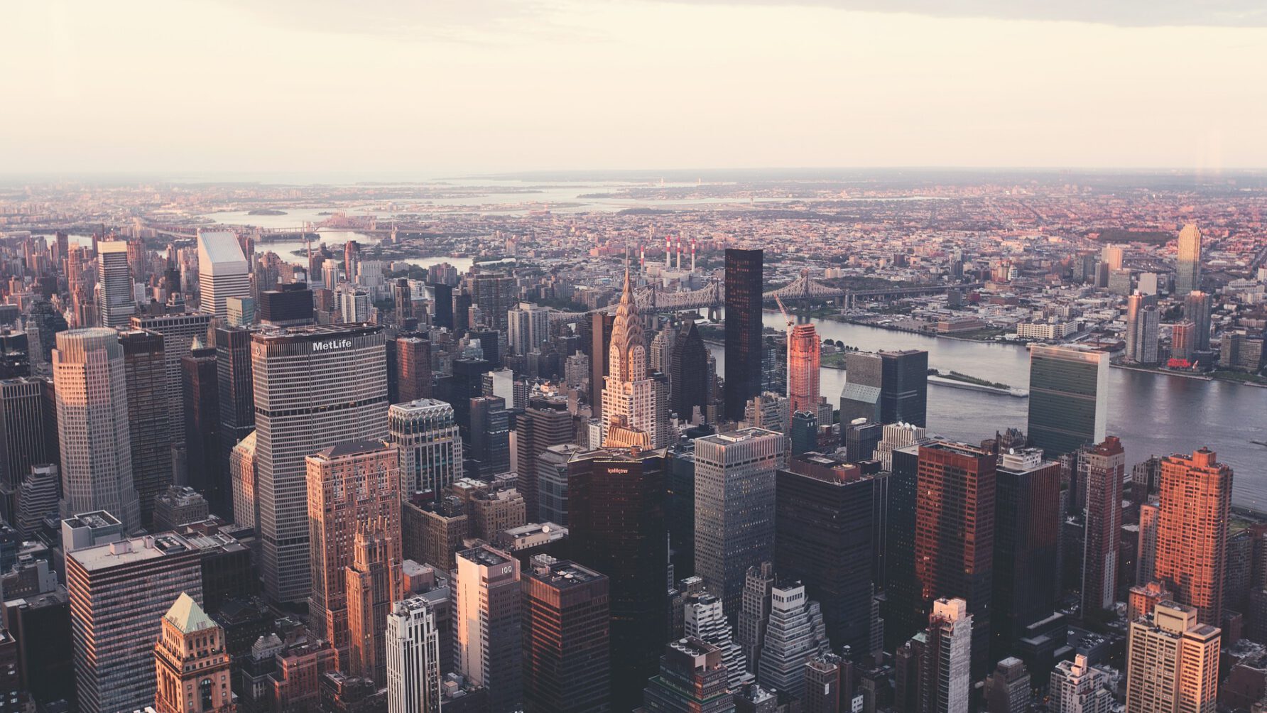 An expansive aerial view of the New York City skyline during sunset, showing the Chrysler Building and other Manhattan skyscrapers bathed in warm light, overlooking the East River and distant bridges.