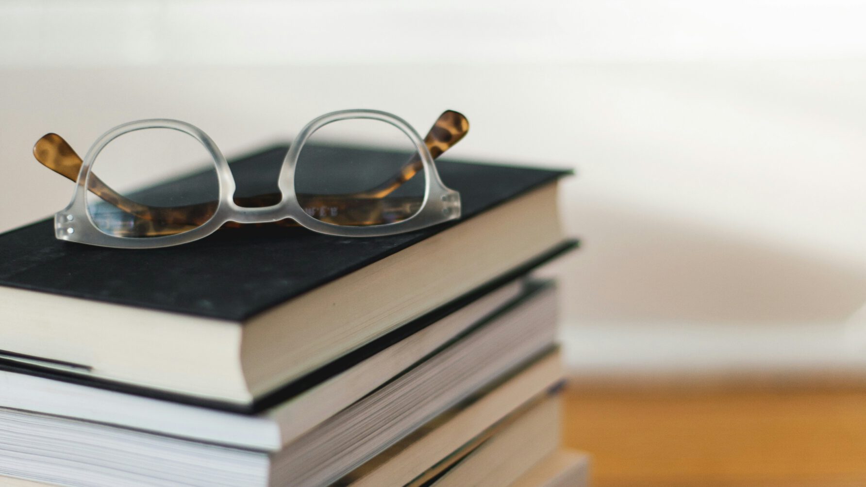 A pair of translucent frames with tortoiseshell arms resting on top of a neat stack of several hardcover and paperback books on a wooden surface.