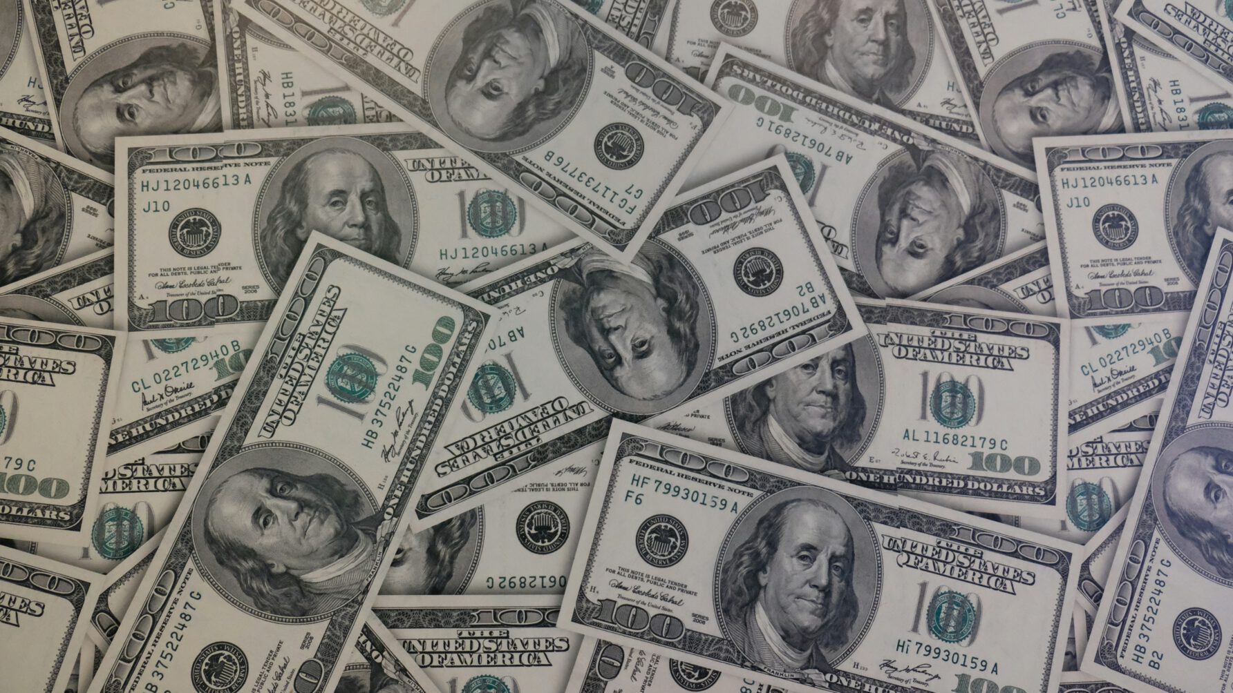 A high-angle, full-frame shot of numerous United States one hundred dollar bills overlapping and scattered across a surface, featuring the portrait of Benjamin Franklin on each note.