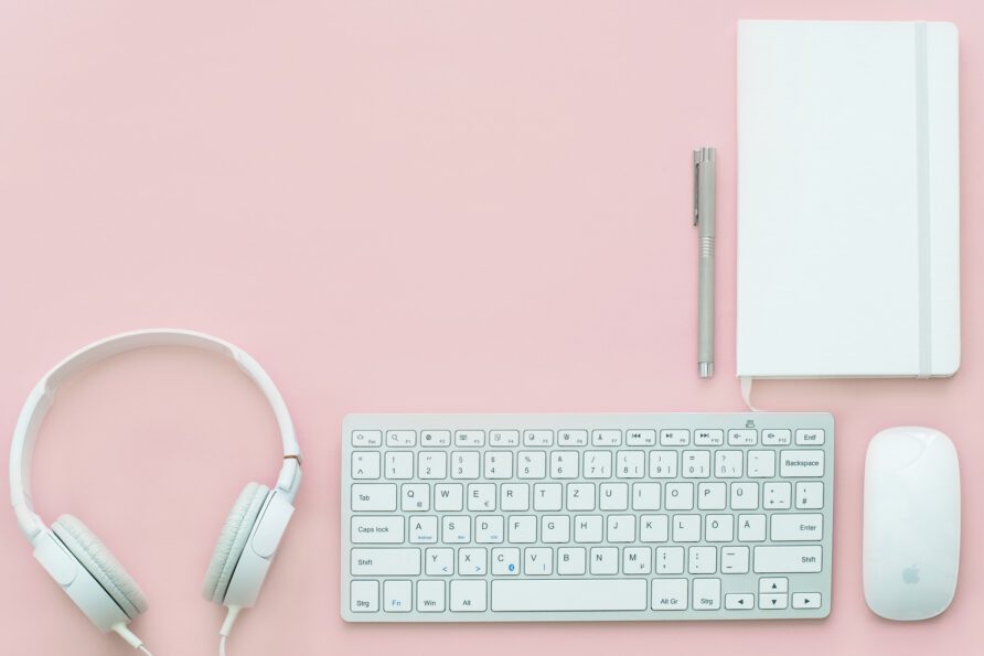 A top-down flat lay view featuring a white keyboard, headphones, a computer mouse, a silver pen, and a closed white notebook arranged on a solid pink surface.